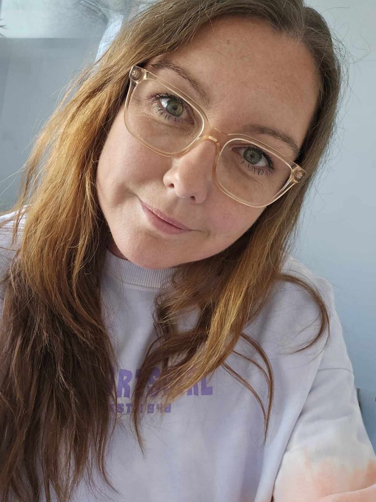 A photo of Bonnie Starick, an Aboriginal woman looking into the camera with clear spectacles and long honey blonde hair falling around her shoulders. She is wearing a white tshirt and is smiling.